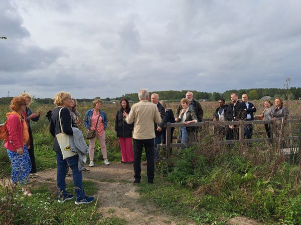 Een groep mensen staat op een pad in een open veld, luisterend naar een spreker die voor hen staat. De groep bestaat uit zowel mannen als vrouwen van verschillende leeftijden, gekleed in casual kleding. Op de achtergrond zijn bomen en een bewolkte lucht zichtbaar.
